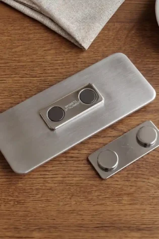The back of a rectangular, brushed metal name tag, featuring a built-in magnetic attachment mechanism with two embedded dark grey magnets. The text 'MAGNETIC ATTACHMENT' is etched on the small frame below the magnets. A matching separate magnetic backing plate with two protruding caps rests next to it on a dark wooden table. Parts of a textured beige linen napkin and the edge of a white ceramic saucer are visible at the top.