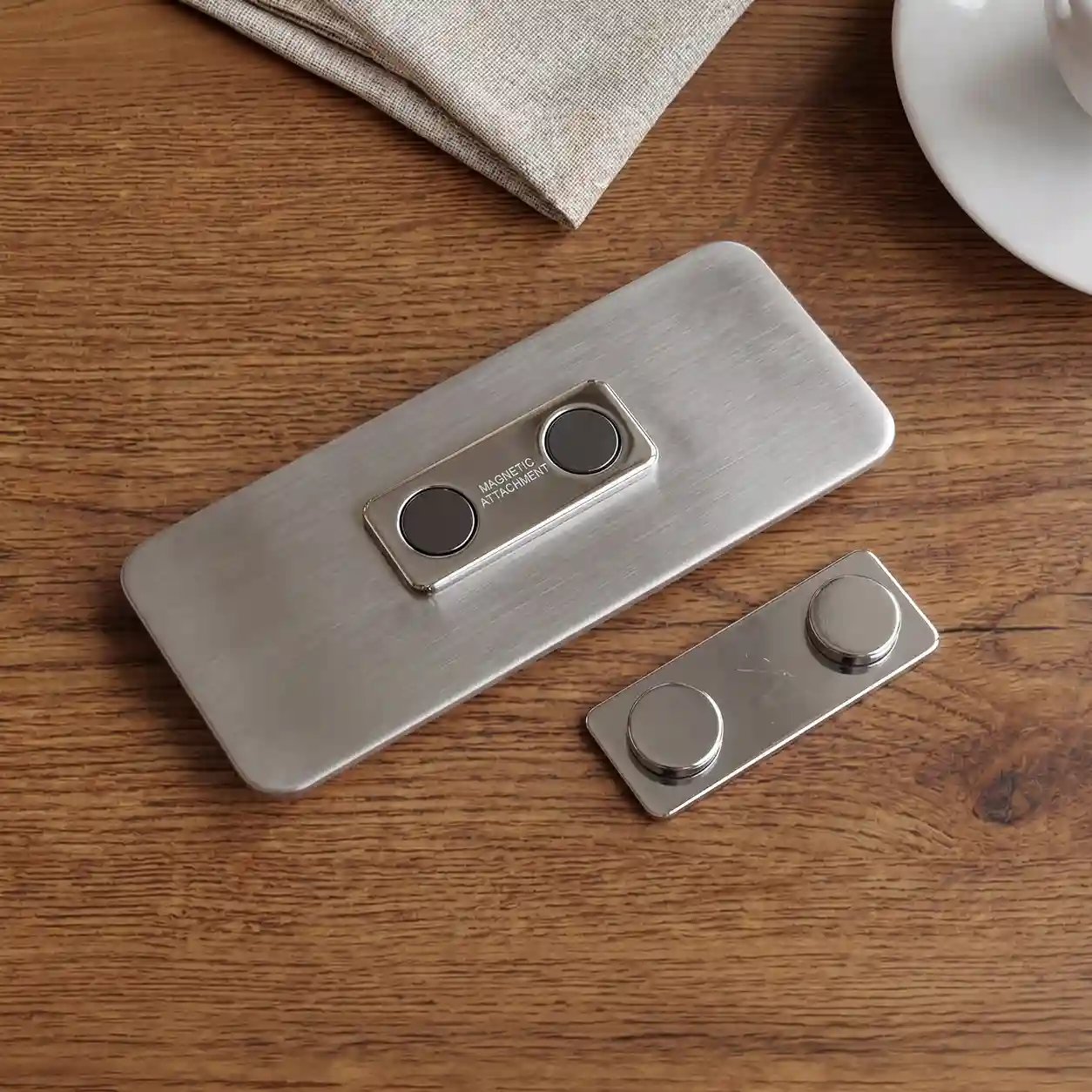 Badge-with-magnet The back of a rectangular, brushed metal name tag, featuring a built-in magnetic attachment mechanism with two embedded dark grey magnets. The text 'MAGNETIC ATTACHMENT' is etched on the small frame below the magnets. A matching separate magnetic backing plate with two protruding caps rests next to it on a dark wooden table. Parts of a textured beige linen napkin and the edge of a white ceramic saucer are visible at the top.