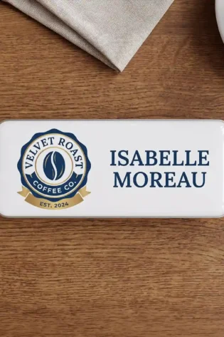 The front of the same rectangular name tag with a polished white domed faceplate, lying centered on the dark wooden table. On the left is the 'VELVET ROAST COFFEE CO.' logo, a dark blue circular badge with a stylized single coffee bean in the center, and a gold banner below reading 'EST. 2024'. To the right, the name 'ISABELLE MOREAU' is printed in large dark blue letters. Parts of the beige napkin and white saucer are visible at the top.