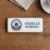 The front of the same rectangular name tag with a polished white domed faceplate, lying centered on the dark wooden table. On the left is the 'VELVET ROAST COFFEE CO.' logo, a dark blue circular badge with a stylized single coffee bean in the center, and a gold banner below reading 'EST. 2024'. To the right, the name 'ISABELLE MOREAU' is printed in large dark blue letters. Parts of the beige napkin and white saucer are visible at the top.