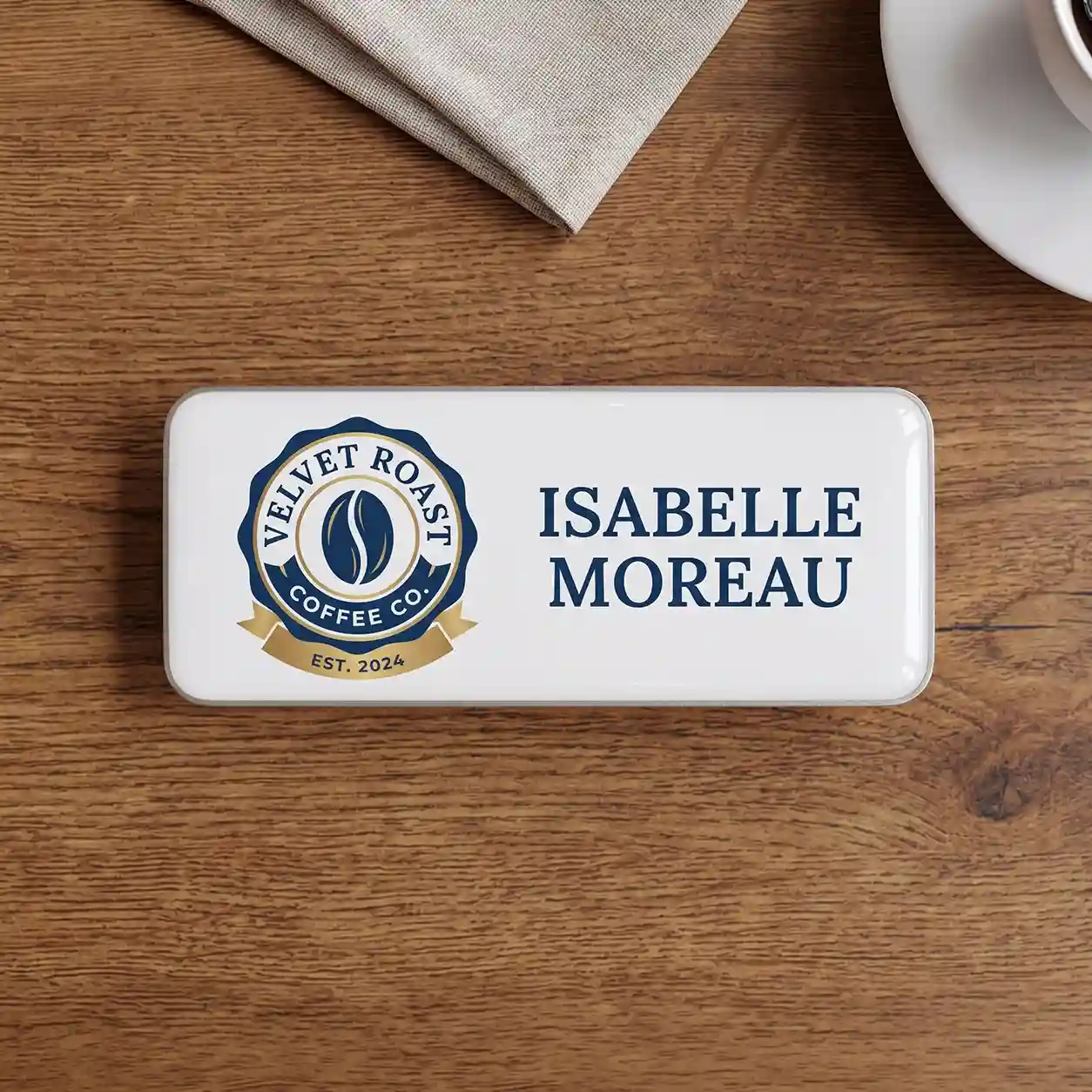 badge-epoxy The front of the same rectangular name tag with a polished white domed faceplate, lying centered on the dark wooden table. On the left is the 'VELVET ROAST COFFEE CO.' logo, a dark blue circular badge with a stylized single coffee bean in the center, and a gold banner below reading 'EST. 2024'. To the right, the name 'ISABELLE MOREAU' is printed in large dark blue letters. Parts of the beige napkin and white saucer are visible at the top.